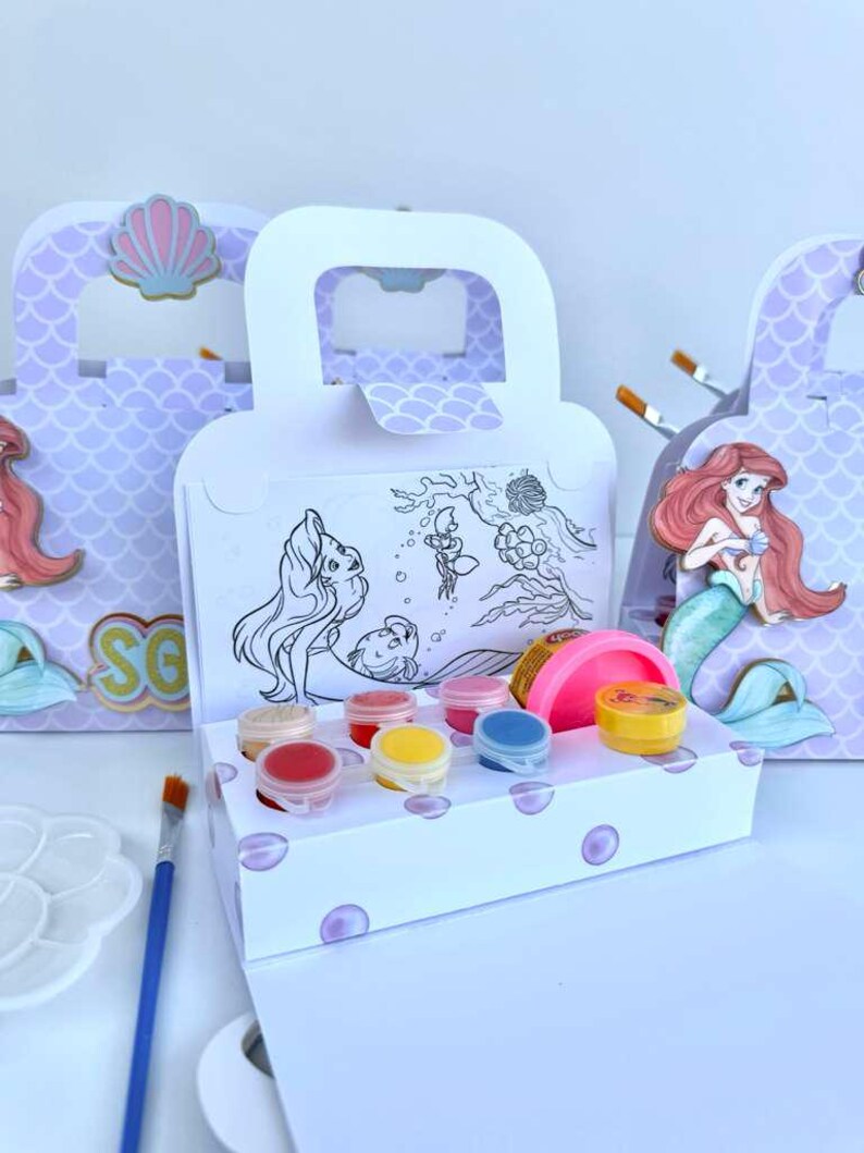 May include: A white cardboard craft box with a handle, featuring a mermaid illustration and a black and white coloring scene. The box contains small circular paint pots in red, yellow, blue, and pink, alongside a pink container of modeling dough.