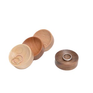 May include: A set of four wooden ring dishes, three are light wood and one is dark wood. The dishes are stacked on top of each other. Two rings are in one of the dishes.