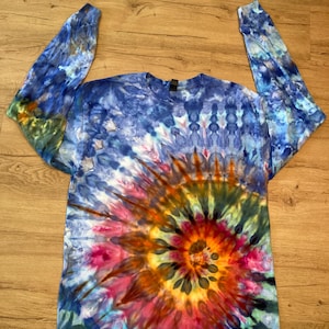 May include: A long-sleeved crew neck shirt featuring a vibrant tie-dye pattern. The design showcases a central sunburst motif in shades of orange, yellow, pink, and deep red, surrounded by a swirling background of blue, purple, and white hues.