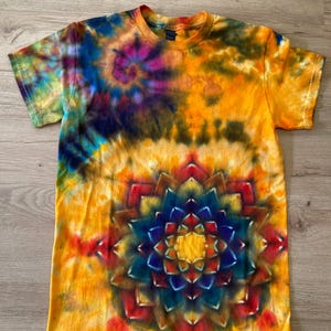 May include: A short-sleeved tie-dye t-shirt with a vibrant, colourful design. The shirt features a large, symmetrical mandala pattern in the centre, with shades of blue, red, and yellow. The background is a warm yellow, with swirls of blue, purple, and green.