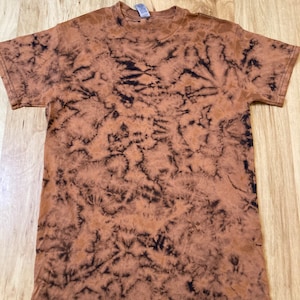 May include: A brown t-shirt with a tie-dye pattern. The shirt has a crew neck and short sleeves. The tie-dye design features dark brown and black patterns against a lighter brown background. The shirt is made of a soft material.