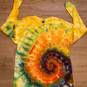 May include: A long-sleeved tie-dye shirt with a spiral pattern. The shirt displays a vibrant mix of yellow, orange, green, and purple. The sleeves are extended, showing the full length of the garment. The shirt is made of a soft material.