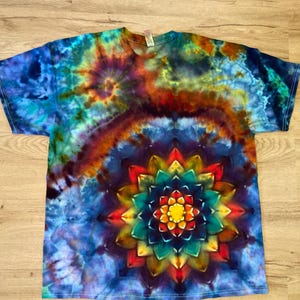 May include: A tie-dye t-shirt with a vibrant, colorful design. The shirt features a large, symmetrical floral pattern in the center, with a spiral and swirling pattern in shades of blue, orange, yellow, and green.