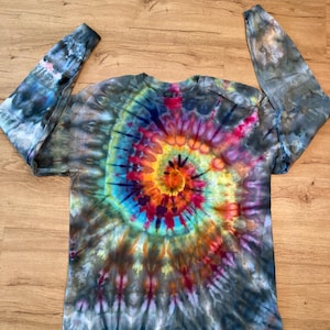 May include: A long-sleeve crew neck shirt featuring a vibrant tie-dye spiral pattern. The design radiates from a central point in shades of bright orange, yellow, and magenta, transitioning into deep blues, purples, and dark charcoal gray tones.