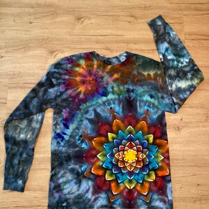May include: A long-sleeve shirt featuring a vibrant tie-dye pattern with a large, multi-coloured mandala design in shades of yellow, orange, red, and blue. The background consists of dark, smoky swirls of black, grey, and deep blue tones.