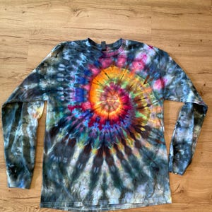 May include: A long-sleeve crew neck shirt featuring a vibrant tie-dye spiral pattern. The center of the spiral displays bright orange, yellow, and red hues, which transition into deep blues, purples, and dark charcoal tones across the fabric.
