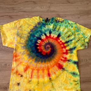 Puede incluir: Una camiseta amarilla teñida con un diseño en espiral. La espiral presenta una vibrante mezcla de colores, incluyendo rojo, naranja, azul y verde. La camiseta está hecha de tela suave y tiene un cuello redondo clásico.