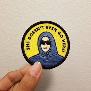 Puede incluir: Un parche bordado amarillo y azul con un personaje de dibujos animados que lleva una sudadera azul y gafas de sol. El texto en el parche dice "She Doesn't Even Go Here!"