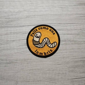 以下が含まれることがあります： 笑顔のミミズと黒い文字「Feliz como una lombriz」が刺繍された黄色のパッチ