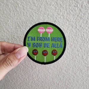 Pode incluir: Etiqueta bordada verde com o texto "I'm from here y soy de allá" e três pirulitos com rostos marrons e lábios vermelhos.