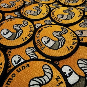以下が含まれることがあります： 黄色い背景と黒い輪郭を持つ刺繍パッチのクローズアップ。パッチには、漫画のミミズが描かれており、「Feliz como una lombriz」というテキストが書かれています。これは、「ミミズのように幸せ」という意味です。