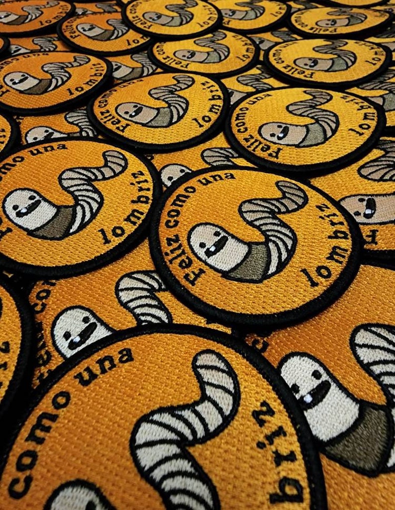 以下が含まれることがあります： 黄色い背景と黒い輪郭を持つ刺繍パッチのクローズアップ。パッチには、漫画のミミズが描かれており、「Feliz como una lombriz」というテキストが書かれています。これは、「ミミズのように幸せ」という意味です。