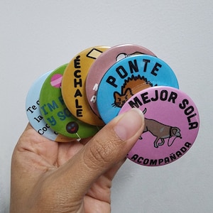 May include: A hand holds a fan of colorful round buttons. Each button features a unique design and text, including a dog, a cat, and various phrases in Spanish. The buttons are in shades of blue, green, yellow, pink, and brown.
