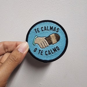 May include: A blue and black embroidered patch with the text "Te Calmas o Te Calmo" and a cartoon image of two hands shaking.