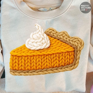 May include: Cream-coloured sweatshirt featuring a knitted pumpkin pie slice with whipped cream. The pie has a golden-yellow filling and a braided crust. The design is centered on the chest.