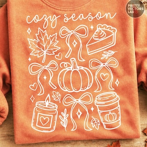 May include: An orange sweatshirt featuring white line art graphics of a maple leaf, bows, a slice of pie, a pumpkin, a candle, and a coffee cup. The text "cozy season" is printed in white script at the top. A circular logo is in the top right corner.