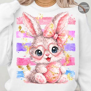 Może przedstawiać: Biała bluza z ilustracją królika. Królik ma okulary w kształcie serca i kokardę, trzymając różowe jajko. Projekt zawiera różowe, fioletowe i niebieskie paski ze złotymi akcentami, idealny na Wielkanoc.