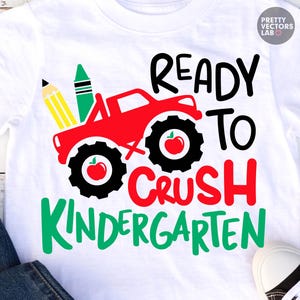 Puede incluir: Una camiseta blanca con un gr&aacute;fico de un cami&oacute;n monstruo rojo con neum&aacute;ticos negros, un l&aacute;piz amarillo y un cray&oacute;n verde. El dise&ntilde;o incluye manzanas rojas dentro de los neum&aacute;ticos y el texto READY TO CRUSH KINDERGARTEN en negro, rojo y verde.