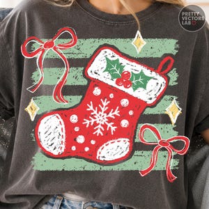 Könnte beinhalten: Ein anthrazitfarbenes T-Shirt mit einer Grafik eines roten Weihnachtsstrumpfes, verziert mit einer weißen Schneeflocke, weißen Punkten und einem weißen Umschlag mit grünen Stechpalmenblättern und roten Beeren, akzentuiert durch rote Schleifen.
