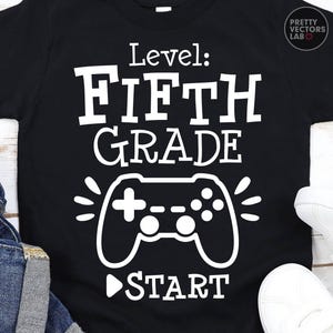 Könnte beinhalten: Ein schwarzes kurzärmeliges T-Shirt mit weißem Siebdrucktext, der Level: FIFTH GRADE und START lautet, begleitet von einer Grafik eines Videospiel-Controllers. Das Design ist mittig auf der Vorderseite des Kleidungsstücks platziert.