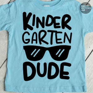 May include: A light blue short-sleeved t-shirt featuring bold black text that reads KINDERGARTEN DUDE. The design includes a graphic of black sunglasses positioned between the words KINDERGARTEN and DUDE, centered on the front of the shirt.