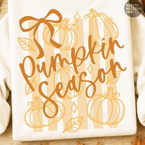 Puede incluir: Una sudadera color crema con un estampado gráfico que incluye el texto Pumpkin Season en cursiva naranja. El diseño presenta dibujos de calabazas en líneas naranjas, un lazo, hojas y pequeñas estrellas sobre rayas verticales beige.