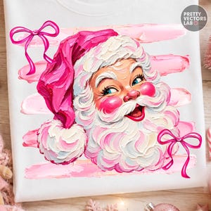 Puede incluir: Una camiseta blanca con un gráfico de Santa Claus con gorro y barba rosa, realizado en un estilo de pintura al óleo con relieve. El diseño incluye rayas de pinceladas rosas de fondo y dos lazos rosas, con el texto Pretty Vectors Lab.