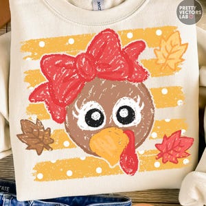 May include: A cream-colored crewneck sweatshirt featuring a graphic of a brown turkey face with a large red bow, white polka dots, and yellow stripes. The design includes three autumn leaves in shades of brown, yellow, and red, with a distressed texture.