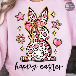 Może przedstawiać: Jasnoróżowa bluza z nadrukiem wielkanocnego królika w panterkę, różową kokardą, gwiazdkami i sercami. Słowa "happy easter" są napisane czarną czcionką pod królikiem. Projekt ma złote akcenty.