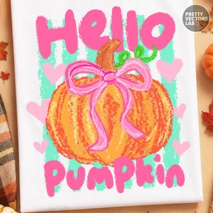 Puede incluir: Una camiseta blanca con un gráfico de una calabaza naranja con un lazo rosa, rodeada de corazones rosas y rayas verticales de color verde azulado. El texto "Hello Pumpkin" está impreso en letras rosas grandes sobre y bajo la calabaza.