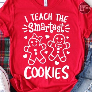 May include: A red short-sleeved t-shirt featuring white graphic text that reads I TEACH THE SMARTEST COOKIES. The design includes two gingerbread figures, one with a bow and one with a bowtie, surrounded by small white heart shapes.