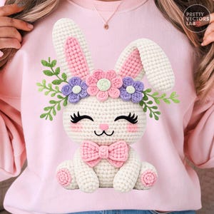 Puede incluir: Una sudadera rosa claro con un diseño de conejito tejido a ganchillo. El conejito tiene pelaje blanco, detalles rosas en las orejas y mejillas, y una pajarita rosa. Está adornado con una corona floral de flores rosas y moradas y hojas verdes.