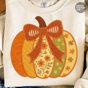 Op de afbeelding: Crèmekleurige sweatshirt met een applicatie van een pompoen. De pompoen heeft oranje, gele en groene secties, een bruine steel en een decoratieve strik. De strik heeft een gestreept patroon en bloemendetails. De tekst "PRETTY VECTORS LAB" is zichtbaar.