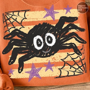 May include: An orange sweatshirt featuring a graphic print of a black spider with large white eyes and a small smile. The design includes black spiderwebs in the corners, purple stars, and horizontal beige stripes. The text PRETTY VECTORS LAB is visible.