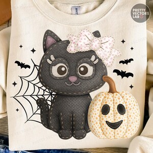 Op de afbeelding: Crèmekleurige sweatshirt met een zwarte kat met een roze strik, een lachende pompoen, een spinnenweb, vleermuizen en sterren. Het ontwerp heeft een gestikte uitstraling, perfect voor Halloween.