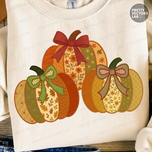 以下が含まれることがあります： クリーム色のスウェットシャツ。模様入りのセグメントとリボンが付いた装飾的なカボチャが3つ描かれています。カボチャはオレンジ、緑、黄色の色合いで、花柄と葉柄があります。リボンはコーディネートされた色です。スウェットシャツはクルーネックです。