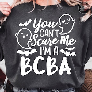 Op de afbeelding: Donkergrijze sweatshirt met witte tekst en afbeeldingen. De tekst luidt "You Can't Scare Me I'm a BCBA" met spook- en vleermuisafbeeldingen. De sweatshirt heeft een ronde hals.