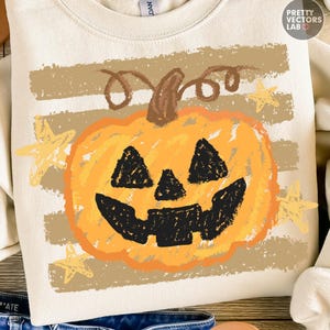 May include: A cream-colored crewneck sweatshirt featuring a graphic of an orange jack-o'-lantern pumpkin with a brown stem. The design includes black triangular eyes, a nose, and a jagged mouth, set against horizontal tan stripes and yellow stars.