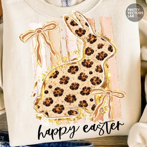 Könnte beinhalten: Cremefarbenes Sweatshirt mit einem Leopardenmuster-Hasen-Design mit goldenen Akzenten und Schleifen. Die Worte "happy easter" sind in schwarzer Schrift unter dem Hasen geschrieben. Das Design umfasst rosa vertikale Streifen.