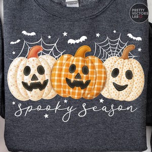 Puede incluir: Sudadera gris oscuro con tres calabazas aplicadas con caras de jack-o'-lantern. Las calabazas son en tonos naranja y crema, con una telaraña y murciélagos. El texto "spooky season" está escrito en una fuente de guión.