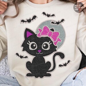 May include: A cream sweatshirt with a black cat design, a pink bow, and a purple ear. The cat is set against a grey moon, with bats flying around. The design is a cute, Halloween-themed graphic.