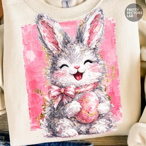 May include: Cream-colored sweatshirt featuring a smiling bunny illustration. The bunny is gray with pink accents, holding a pink egg, and wearing a pink bow. The background is a textured pink rectangle with gold flecks. The text "PRETTY VECTORS LAB" is in the upper right corner.