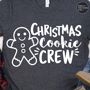 May include: A dark heather gray short-sleeve t-shirt featuring a white graphic of a gingerbread person and the text "CHRISTMAS Cookie CREW" in a mix of block and script fonts. The shirt has a round neckline and a small brand tag at the collar.