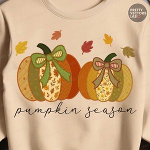 Op de afbeelding: Beige sweatshirt met twee decoratieve pompoenen met groene strikken en herfstbladaccenten. De pompoenen hebben patchwork-ontwerpen in tinten oranje, groen en geel. De tekst "pumpkin season" is geschreven in een cursief lettertype.