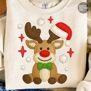 May include: Cream-colored sweatshirt featuring a reindeer design with a red nose, brown antlers, and a green bow tie. The reindeer wears a red and white Santa hat. Red stars and white snowflakes surround the reindeer. The text "PRETTY VECTORS LAB" is visible.