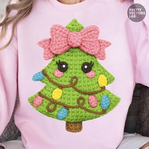 May include: A pale pink sweatshirt with a crocheted Christmas tree design. The green tree has a pink bow, colourful lights, and cute eyes. The design is centered on the front of the sweatshirt. The text "PRETTY VECTORS LAB" is in the upper right corner.