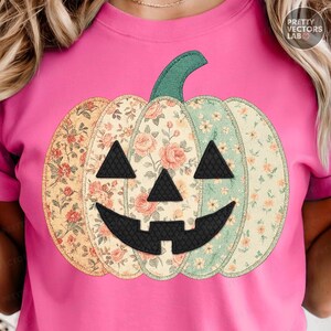 Op de afbeelding: Roze T-shirt met een patchwork pompoen ontwerp. De pompoen heeft een groene stengel en een zwart jack-o'-lantern gezicht. De pompoen secties zijn bewerkt met bloemenprints in tinten oranje, roze en groen.