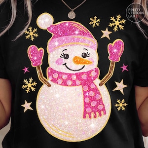 May include: Black t-shirt featuring a glittery snowman design. The snowman wears a pink hat, scarf, and mittens, with a carrot nose and a smiling face. Gold snowflakes and stars surround the snowman, creating a festive winter theme.