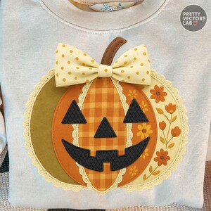 May include: Cream-colored sweatshirt featuring a large, appliqued pumpkin design. The pumpkin has a plaid center, black facial features, and a yellow bow. The design is surrounded by a scalloped edge and floral accents, perfect for fall.