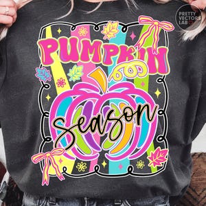 May include: Dark gray sweatshirt featuring a colorful graphic design. The design includes the words "PUMPKIN SEASON" in bright pink and teal, with a pumpkin illustration and decorative elements like leaves and bows.